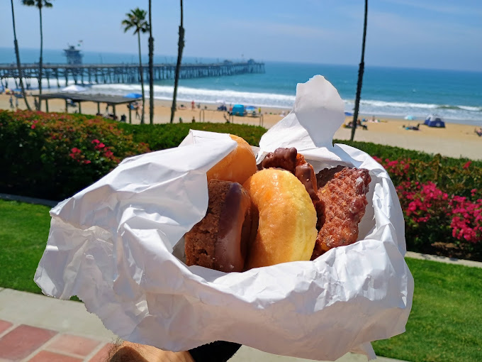 A bag of fresh donuts and pastries from Donut House, with the San Clemente pier and beach in the background